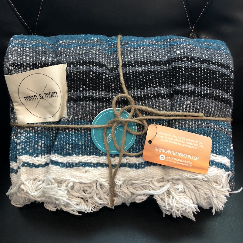 The MNTN & MOON Lago Mexican Falsa Blanket came wrapped in reusable twine and measures 48” wide x 73” long. Made of 50% acrylic, 10% cotton, and 40% polyester, it’s very soft. The blankets are handwoven by artisans in Mexico and hand finished in the United States.
